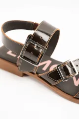Sandalias de cuero charolado negro con dos tiras con hebillas metálicas plateadas y tira en el talón. Suela plana color marrón.