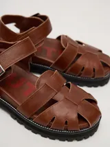 Sandalias de cuero marrón con tiras entrecruzadas, suela de goma negra con pespuntes blancos y hebilla lateral.