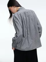 Camisa de corte clásico con estampado de cuadros vichy en tonos azul y blanco. Presenta cuello camisero, cierre frontal con botones ocultos y mangas largas con puños abotonados. Incluye un detalle de broche metálico dorado en el cuello.