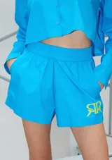 Short de poplín de algodón color azul vibrante, con cintura elástica ancha, bolsillos laterales y estampa de logo en color amarillo neón en el lateral izquierdo.