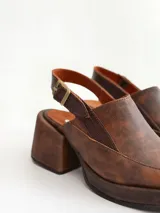 Zapato tipo zueco de cuero color chocolate con efecto desgastado, plataforma y taco cuadrado. Presenta una tira trasera ajustable con hebilla metálica.