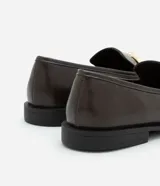 Zapato mocasín femenino de PU color negro, con punta cuadrada y detalle metálico dorado en el empeine. Tiene un tacón bajo y suela de goma.