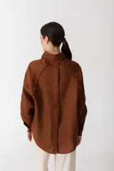 Camisa color ladrillo con rayas verticales finas color crema, de corte recto y mangas largas con puños abotonados. Presenta cuello camisero, cierre frontal con botones y detalle de flecos en los hombros.