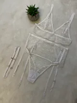 Conjunto de lencería de encaje blanco compuesto por un bralette triangular, una tanga con tiras laterales y un portaligas integrado. El diseño presenta detalles de encaje floral y breteles regulables.