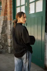 Camisa de lino color negro con silueta relajada, cuello camisero y cierre frontal con botones de nácar. Presenta mangas largas con puños y detalles de tiras laterales.