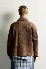 Cazadora de piel con acabado en ante color marrón, de corte relaxed fit, con cuello solapa, mangas largas con puños abotonados, bolsillos de vivo en pecho y cadera, y cierre frontal con cremallera oculta por solapa con botones a presión.