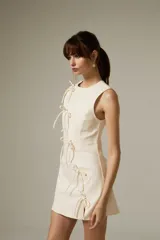 Vestido corto de jean color natural, con corte al cuerpo, cuello a la base y diseño sin mangas. Presenta un detalle de cierre frontal con lazos decorativos y una pequeña abertura lateral.