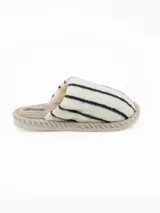 Pantuflas abiertas de estilo mule, con capellada de peluche color crema con rayas negras horizontales y plantilla de pana acanalada en tono beige.