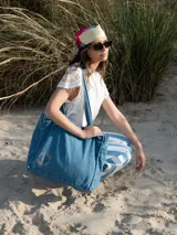 Bolso tipo tote confeccionado en tela denim de color azul claro. Presenta un diseño amplio con costuras visibles, doble asa de hombro y un detalle bordado circular en color rojo en la parte inferior.