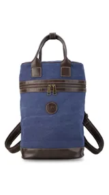 Mochila matera de diseño vertical, confeccionada en tela canvas marrón con detalles y refuerzos en cuero sintético oscuro. Cuenta con doble asa superior de mano, correas traseras regulables, cierre metálico frontal y bolsillo posterior.