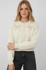 Cardigan de punto color natural con cuello redondo y cierre frontal de botones. Presenta un delicado bordado de lazos en la parte superior del pecho y puños acanalados.