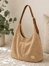 Cartera tipo hobo de gamuza color avena con diseño amplio y costuras decorativas. Cuenta con correa ajustable de cuero marrón y una placa metálica dorada con el logo de la marca en la parte inferior.