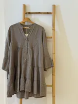 Vestido amplio de lino color marrón topo, con cuello mao y abertura frontal con botones de coco. Presenta mangas cortas con volados y un diseño escalonado con frunces en el cuerpo.