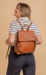 Mochila tipo cartera con tapa, confeccionada en símil cuero color suela. Cuenta con doble compartimento, división interna, cierre de seguridad y bolsillo trasero con cierre. Incluye una correa frontal decorativa con tachas metálicas.