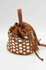 Cartera tipo bucket bag de gamuza negra, decorada con múltiples tachas metálicas plateadas de forma circular. Cuenta con cierre de cordón ajustable y asa superior.