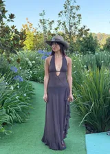 Vestido largo de estilo etéreo y fluido, color marrón, con escote halter profundo y diseño cut-out en la cintura. La falda presenta un detalle de volados laterales y una caída ligera.