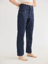 Jean de mezclilla azul oscuro, corte recto y tiro alto, con pespuntes contrastantes en color amarillo verdoso. Presenta detalles de atraques y remaches dorados en los bolsillos delanteros.