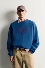 Sudadera azul con corte relaxed fit, cuello redondo y manga larga, confeccionada en tejido de algodón. Presenta un bordado frontal a contraste en color rojo con diseño ornamental y texto.