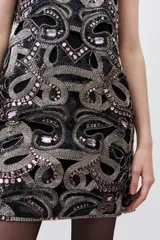 Vestido mini negro sin mangas y cuello redondo, completamente bordado con mostacillas, piedras y lentejuelas en tonos dorados y rosados. Presenta un corte recto con calados que acompañan el diseño.