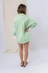 Camisa verde pastel de algodón, corte oversize, con cuello camisero, cierre con botones, mangas largas con puños con volados de encaje blanco, dos bolsillos frontales con solapa y ruedo irregular. 