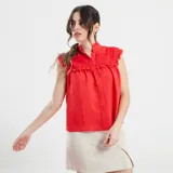 Camisa sin mangas color beige, con cuello alto con volados, detalles de encaje calado en los hombros y parte superior del pecho, y cierre frontal con botones.