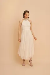 Vestido midi de fiesta color crudo, con cuello halter alto de microtul drapeado, escote cerrado y falda con vuelo y capas de tul.