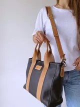 Cartera de cuero negro con detalles en cuero color suela, incluyendo asas fijas y correa larga ajustable. Presenta una etiqueta frontal con logo grabado y cierre superior.