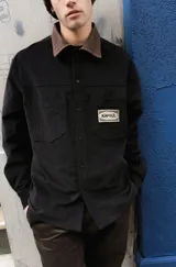 Sobrecamisa negra de corte clásico con cuello camisero en contraste color marrón a cuadros. Presenta cierre frontal con botones, dos bolsillos frontales tipo parche y una etiqueta decorativa con el logo de la marca en el pecho.