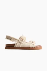 Sandalias planas de sarga de algodón en color beige con terminación desflecada. Cuentan con tiras ajustables con detalle metálico dorado y cierre autoadherente, además de una tira ajustable en el talón. La plantilla es preformada en tono marrón.