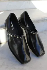 Zapato de cuero vacuno color negro, con punta cuadrada y detalle de tachas metálicas planas en el borde superior. Presenta una costura central vertical y taco geométrico.