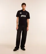 Remera de corte boxy confeccionada en algodón, con cuello polo y cierre frontal de botones. Presenta un diseño estampado en el frente con el número 93, una cruz y la palabra MAD en tipografía gótica.