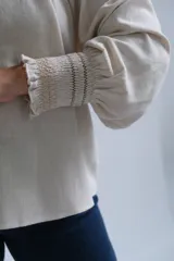 Blusa de corte holgado en color beige, con cuello redondo fruncido y escote en V. Presenta mangas tres cuartos con terminación elástica y un diseño minimalista de textura natural.