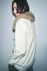 Camisa de popelín color crudo con cuello solapa y aplicación desmontable de pelo sintético en tono marrón. Presenta cierre frontal con botones, manga larga con puños y bajo asimétrico.