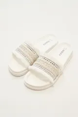 Sandalias de base plana color marfil con tira ancha decorada con bandas de cordón trenzado, detalles plateados con tachas y flecos en los bordes.
