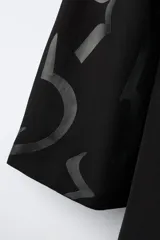 Capa de estilo oriental en color negro, con cuello mao, cierre frontal de alamares y mangas anchas. Presenta un estampado gráfico tonal en toda la prenda y un cinturón de cuerda trenzada ajustable en la cintura.