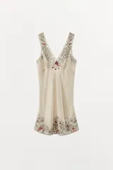 Vestido mini tipo lencero de satén color crema, con escote en pico y tirantes anchos. Presenta bordados florales detallados con aplicación de abalorios, flores y lazos en relieve a contraste en el escote y el ruedo. Cierre en la espalda con lazada.