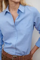 Camisa blanca de manga larga con cuello clásico y botones frontales. Tiene un bolsillo en el pecho con un pequeño logo bordado.