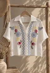 Camisa beige de manga corta con cierre frontal de botones, escote en V y bordados artesanales de flores en tonos azules, amarillos y rojos, acompañados de patrones geométricos.