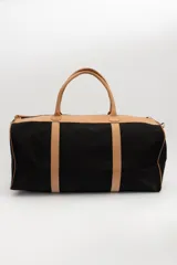 Bolso de viaje tipo duffel de lona negra con detalles en cuero tostado, asas de mano y correa ajustable para el hombro.