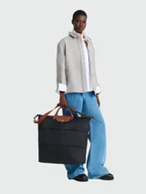 Bolso de viaje Longchamp modelo Le Pliage Original, de diseño ampliable, confeccionado en nylon negro con detalles en cuero marrón en las asas y solapa frontal con cierre de botón a presión. Incluye correa ajustable y extraíble.