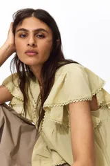 Blusa de tejido liviano color amarillo claro, con escote en V profundo, mangas cortas estilo mariposa y cierre frontal con botones. Presenta detalles de alforzas e inserciones de encaje en el frente y en el borde del ruedo y las mangas, terminando en un volante inferior.