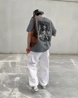 Remera oversize gris oscuro con corte holgado, mangas cortas y estampa trasera en blanco y negro con forma de estampilla que muestra a una persona con un gato y el texto 'Kaboa Industries'.