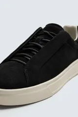 Zapatilla de piel color negro con acabado serraje, cordonera oculta y suela plana a contraste en color crema.