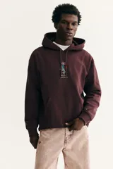 Canguro de corte holgado en color bordó, con capucha ajustable mediante cordones, hombros caídos y bolsillo frontal tipo canguro. Presenta un pequeño estampado central de un jarrón con flores coloridas.