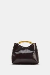 Bolso de mano de efecto acharolado color marrón oscuro, con detalle de tachas metálicas doradas en los bordes. Cuenta con asa rígida metálica dorada y cierre de botón imantado.