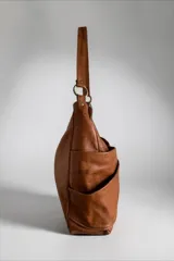 Cartera de cuero natural en color marrón, con diseño envolvente, caída suave y bolsillo exterior cruzado con el nombre de la marca bordado. Cuenta con tiras de hombro, herrajes metálicos y un dije colgante geométrico.