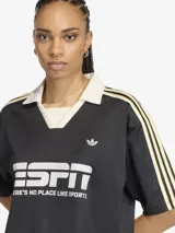 Camiseta de estilo futbolístico con cuello polo, manga corta y corte holgado. Presenta un diseño en color negro con detalles en blanco y amarillo en las mangas, y el logo de la marca junto a un estampado gráfico frontal.