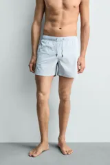 Bañador de hombre de largo medio, color celeste claro con rayas blancas horizontales. Presenta cintura elástica ajustable con cordón, bolsillos frontales y bolsillos traseros de plastrón. Incluye forro interior.
