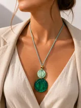 Collar de diseño exclusivo Fahoma, elaborado artesanalmente en Italia. Presenta un colgante compuesto por dos piezas circulares de resina en tonos verde marmolado, unidas por un detalle metálico dorado, suspendidas de un cordón de tela a tono con una cuenta dorada superior.