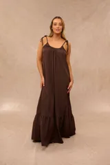 Vestido largo color marrón chocolate, con tirantes ajustables mediante lazos en los hombros, escote redondo fruncido y diseño amplio con volados en la parte inferior.